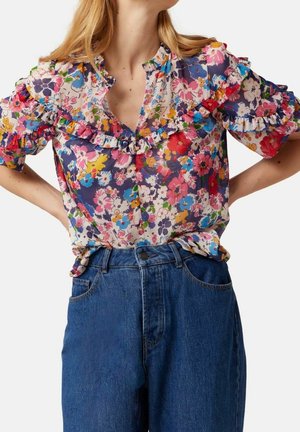 Floral blouse with ruffled collar and sleeves paired with high-waist blue jeans, hands resting on hips, torso visible.