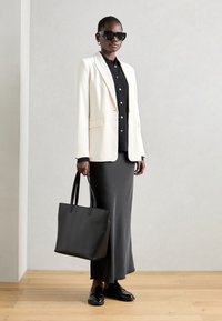 Schwarze Leder-Tote mit zwei Griffen, kombiniert mit einem cremefarbenen Blazer, einem schwarzen Button-Up-Hemd und einem grauen Maxirock. Accessoires umfassen eine Sonnenbrille.