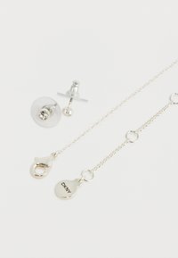 Cercei din argint cu spate clar pentru fixare și o lanț cu carabină. Charm-ul marcat "DKNY" atașat lanțului.