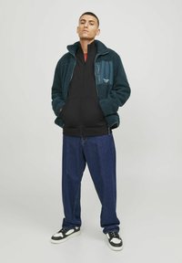Veste en polaire zippée en vert foncé avec un panneau noir, sweat à capuche noir zippé en dessous, jeans bleus amples et baskets noires avec des accents blancs.