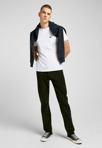 Maglietta bianca con taschino, maglione blu scuro appoggiato sulle spalle, pantaloni verde oliva e sneaker nere con suola bianca.