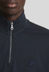 Navyblauwe rits sweatshirt met een hoge kraag. Heeft een gestructureerde stof en een klein geborduurd logo op de linkerborst in bijpassende kleur.