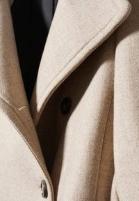 Gros plan sur le revers d'un manteau en laine beige et un bouton noir, mettant en valeur la texture du tissu et les détails de couture sur mesure.