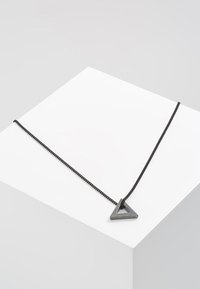 Sort kædelykke med en metallisk, trekantet vedhæng med en struktureret overflade, præsenteret på en hvid geometrisk piedestal.