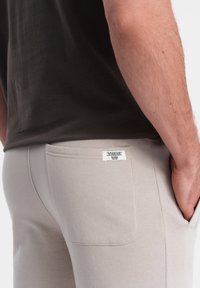 Beige Jogginghose mit glatter Textur, Seitentaschen und einem Etikett auf der Rückseite. Hergestellt aus weichem, dehnbarem Material.