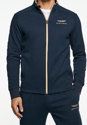 Homme portant un survêtement Aston Martin bleu marine avec une fermeture éclair beige et un logo, debout les mains détendues le long du corps.