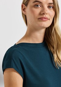 Haut teal à manches courtes avec un encolure large, texture douce et détails de couture subtils au niveau des épaules.