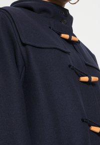 Granatowy wełniany płaszcz z kapturem, z drewnianymi zapięciami na toggle i akcentami w formie lin, o zorganizowanym, dopasowanym kroju.