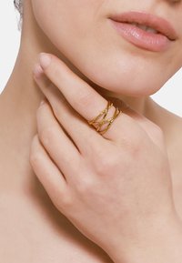 Zlatni uvrnuti prsten nosen na prstima, koji se sastoji od više isprepletenih niti sa teksturiranom površinom. Ton kože je neutralan ispod prstena.