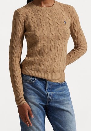 Kvinde iført en beige kabelstrikket sweater med et lille logo på brystet og mellemblå denimjeans.