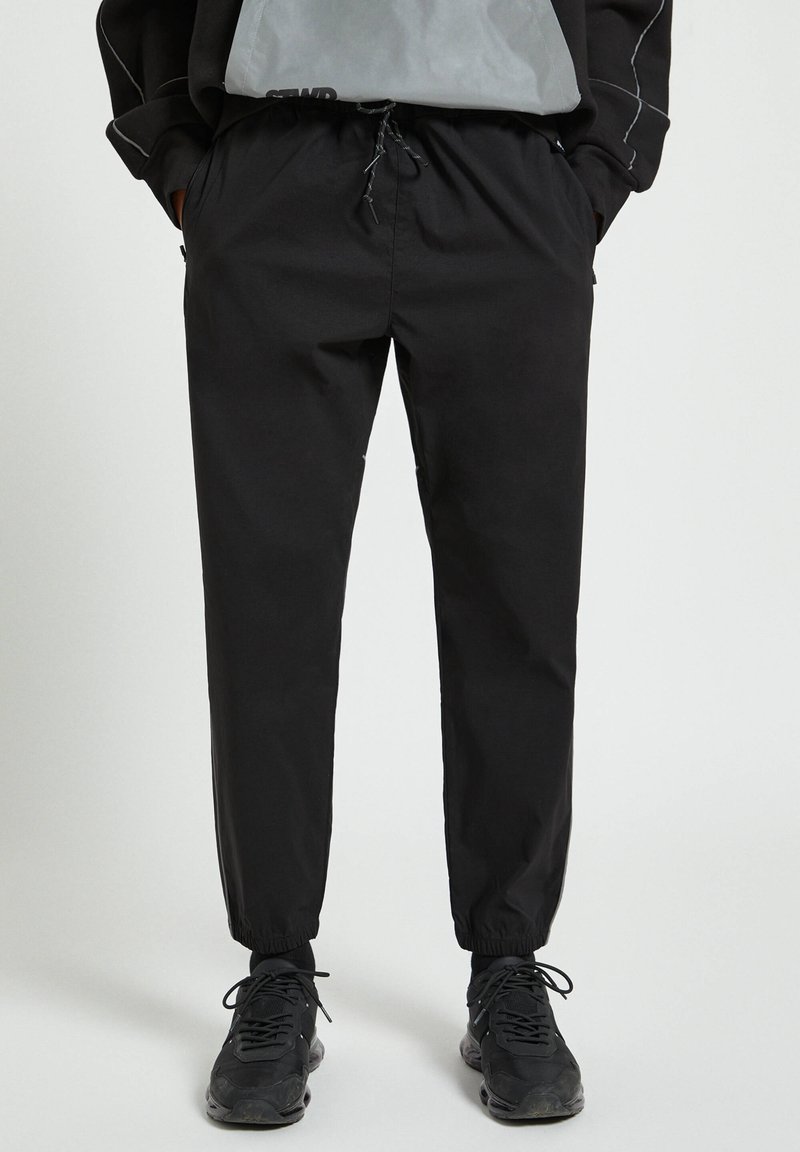 Svarta joggarbyxor tillverkade av lättviktigt tyg, med elastisk midja och snörning, avslutade vid anklarna och sidofickor.