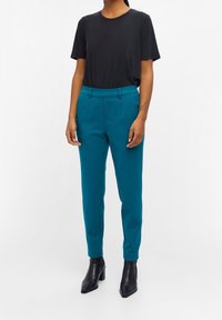 Femme portant un pantalon ajusté bleu sarcelle, un haut noir à manches courtes et des bottines à talons noires, debout devant un fond blanc.
