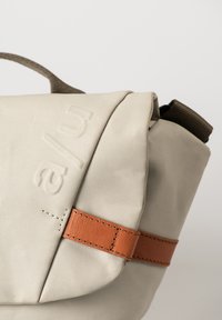 Beige Stofftasche mit runder Form, ausgestattet mit einem braunen Lederakzentgurt und geprägtem Text. Die Oberfläche ist glatt mit sichtbaren Nähten.