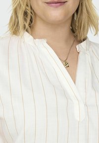 Blusa blanca con finas rayas doradas verticales, con escote en V y tela suave. Un collar de péndulo dorado reposa sobre el escote.