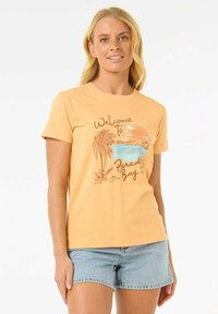 Lichtoranje t-shirt met een grafische afbeelding van een strandscène met palmbomen en de tekst "Welkom in Paradise Bay." Gecombineerd met denimshorts.