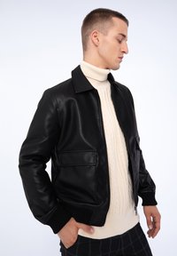 Veste bomber en cuir noir avec fermeture éclair, deux poches avant, poignets côtelés, portée sur un pull en tricot crème à col roulé.