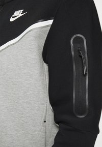 Sudadera deportiva de Nike con bloques de color negro y gris, logo blanco en el pecho y bolsillo con cremallera en la parte superior de la manga negra.