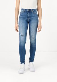 Jeans de mezclilla azul de tiro alto con un ajuste slim, ligero desgaste y detalles desgastados en las rodillas, combinados con zapatillas blancas.