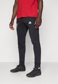Juodos sportinės kelnės su baltu Adidas logotipu ir grafikiniu detalumu, pasižyminčios siauru siluetu ir tekstūruotais kulkšnies rankovėmis, derinamos su juodais batais.