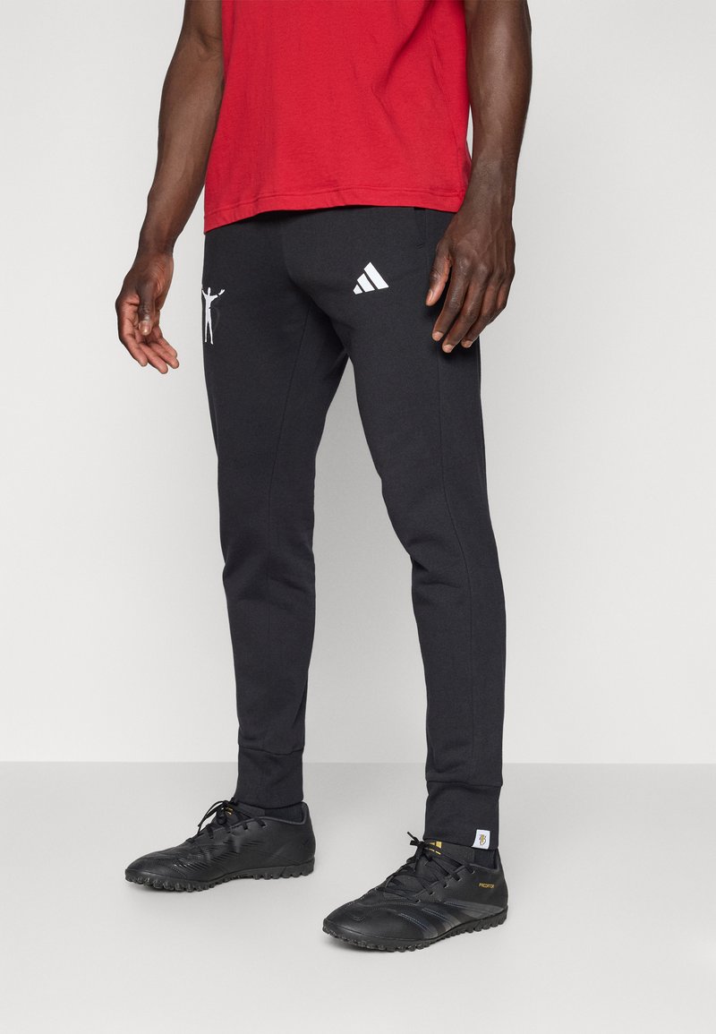 Juodos sportinės kelnės su baltu Adidas logotipu ir grafikiniu detalumu, pasižyminčios siauru siluetu ir tekstūruotais kulkšnies rankovėmis, derinamos su juodais batais.