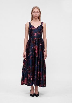 Femme debout portant une robe bleu marine sans manches avec un motif floral rose et des talons noirs sur un fond gris clair uni.