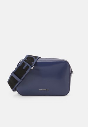 Rechthoekige donkerblauwe crossbodytas met brede zwart-blauwe verstelbare riem en zilverkleurige ritssluiting, "COCCINELLE" logo gecentreerd aan de voorkant.