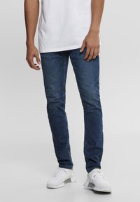 Slim-fit blue denim jeans with a classic five-pocket design, contrasting stitching, and a smooth texture, paired with white sneakers.