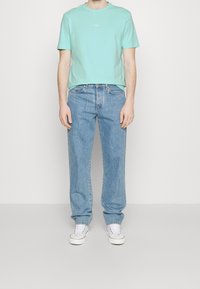 Turkos bomull t-shirt med "BOSS"-logotyp, kombinerad med ljusblå raka jeans. Modellen bär vita sneakers framför en neutral bakgrund.