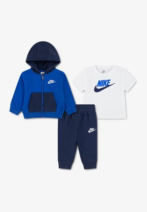 Veste à capuche zippée bleue et marine, t-shirt blanc avec logo bleu, et pantalon de survêtement marine avec logo blanc disposés sur fond blanc.
