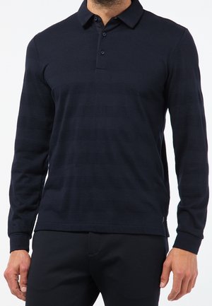 Homme portant un polo noir ajusté à manches longues avec de fines rayures horizontales et un pantalon noir sur fond blanc.