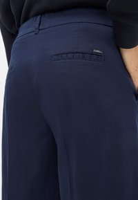 Marineblaue Hose aus glattem Stoff, mit einer Gesäßtasche und einem kleinen metallischen Logo-Acccent. Taillierte Passform mit klaren Linien.
