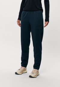 Marineblå joggebukser med elastisk linning, sidelomme og en subtil logo-detalj. Lett stoff med en glatt tekstur.