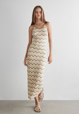 Ung kvinde iført en ærmeløs, ankel-lang kjole med beige og hvide zigzag-mønstre og hvide sandaler mod en ensfarvet baggrund.