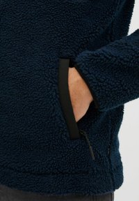 Polarowy sweter w ciemnym granacie, z miękką, teksturowaną powierzchnią, czarnymi wykończeniami wokół kieszeni oraz detalem zamka błyskawicznego u dołu.