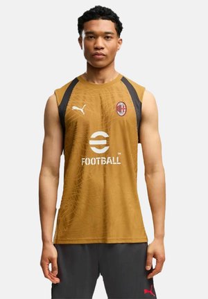 Uomo che indossa una maglia da calcio dorata senza maniche con i loghi Puma e AC Milan e pantaloncini neri con logo Puma rosso, in piedi su sfondo bianco.