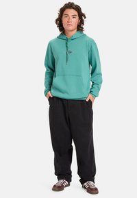 Teal cotton hoodie with a front pouch pocket, adjustable drawstring, and graphic detailing, paired with loose black pants and brown sneakers.