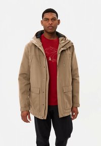 Kaki jas met capuchon en twee voorzakken, met een lichte textuur en contrasterende voering. Gedragen over een rood shirt met een bergafbeelding.