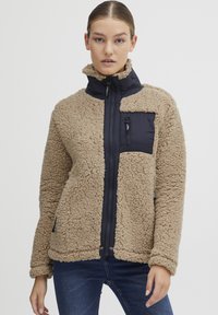 Veste en polaire beige avec un panneau en nylon marine, fermeture éclair complète, col haut et poche poitrine zippée. Texture douce et coupe décontractée.