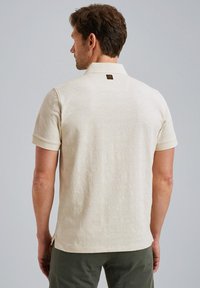 Polo à manches courtes beige en tissu texturé, avec un motif et une petite étiquette de marque au dos du col. Fentes sur les côtés à l'ourlet.