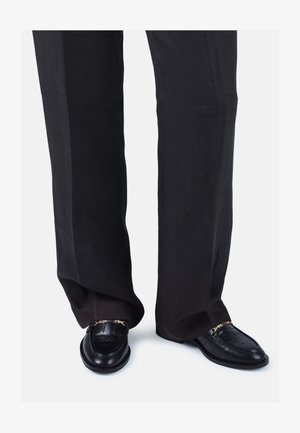 Mustad nahast loafereid krokodillimustriga, kuldne ahelsus, sobitatud avarate tumedate pükstega, näidates laitmatut siluetti.