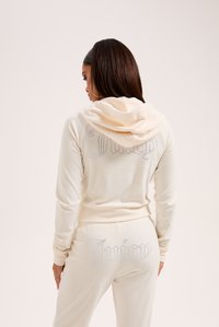 Completo sportivo color crema con un design aderente, dotato di cappuccio e dettagli in strass sulla schiena con la scritta "Juicy" in corsivo.