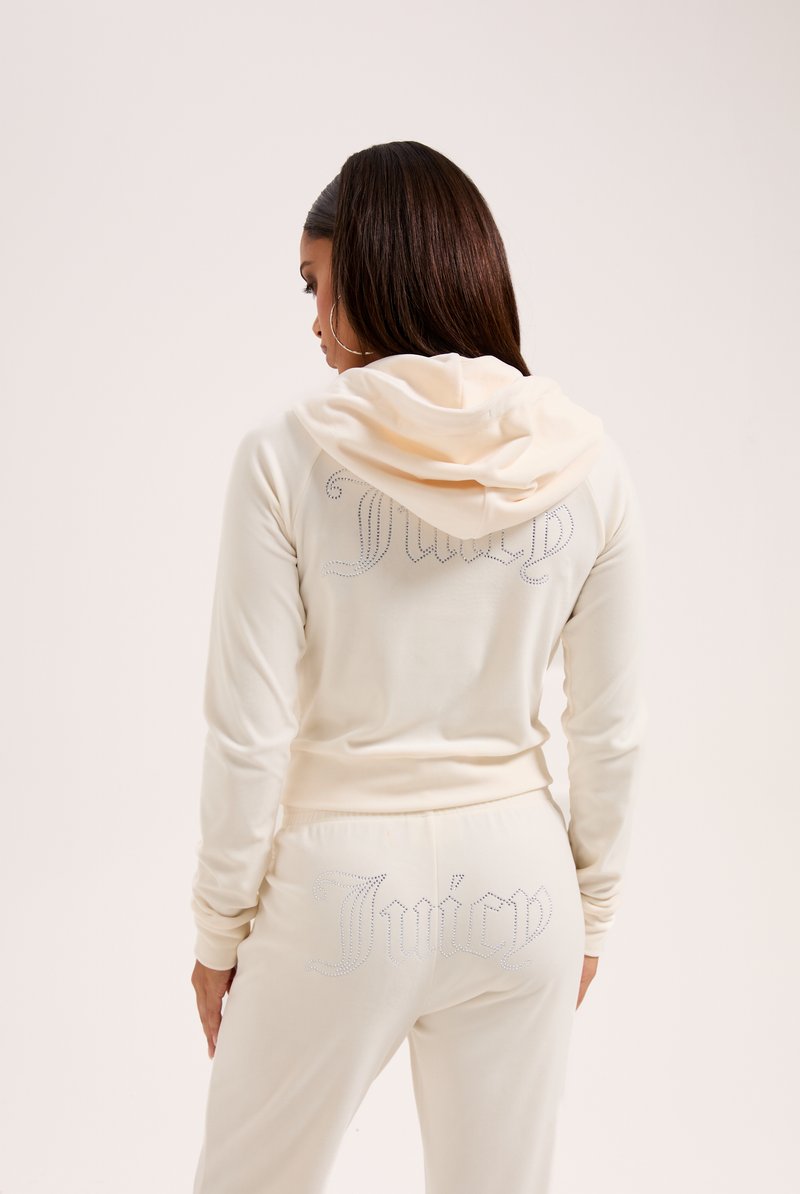 Completo sportivo color crema con un design aderente, dotato di cappuccio e dettagli in strass sulla schiena con la scritta "Juicy" in corsivo.