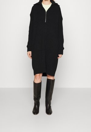 Femme portant une robe noire en maille à manches longues avec une fermeture éclair devant, combinée à des bottes en cuir noires montantes jusqu'aux genoux.