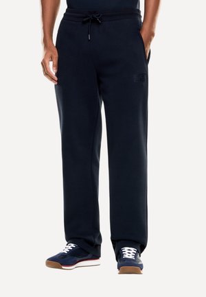 Uomo con pantaloni della tuta neri con coulisse e sneakers blu navy con lacci bianchi, in piedi con le mani in tasca su sfondo bianco.