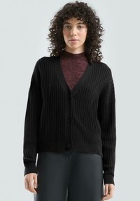 Czarny kardigan w strukturę plastra miodu z dekoltem w szpic, zapinany na guziki, z prążkowanymi mankietami. Nosi się go na teksturowanym bordowym topie. Gładka faktura materiału.