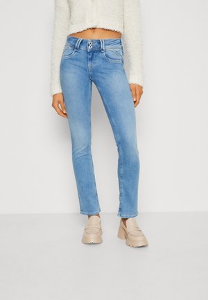 Femme portant un jean bleu clair à double bouton, un pull court pelucheux blanc et des mocassins beiges épais, debout devant un fond uni.