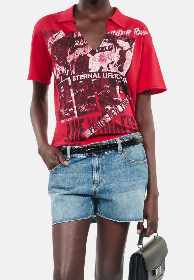 Polo rouge à manches courtes avec un imprimé graphique, présentant un décolleté en V profond et un texte noir. Associé à un short en jean bleu clair et une ceinture noire.