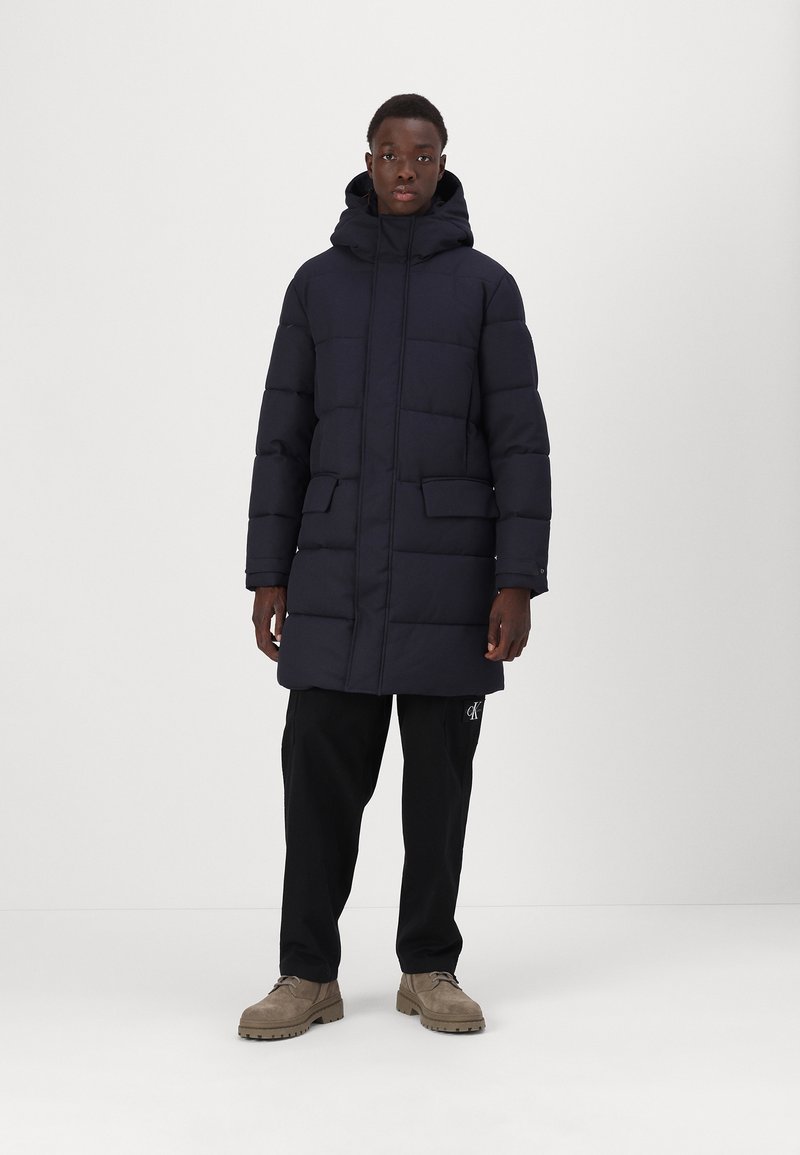 Mørkeblå polstret parka med hætte, der har to forlommer og lynlåslukning. Båret med sorte bukser og beige sko.