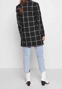 Femme portant un long manteau à carreaux noir et blanc, un jean bleu clair et des bottines blanches à talons, se tenant devant un fond uni.