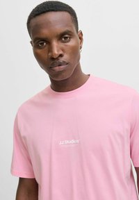 Camiseta de algodón rosa con mangas cortas, que presenta un logo blanco "JJ Studios" en el área del pecho izquierdo, textura suave y corte relajado.